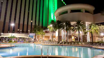 A hotel pool area with lounge chairs, palm trees, and a large building with vertical lines and green lighting.