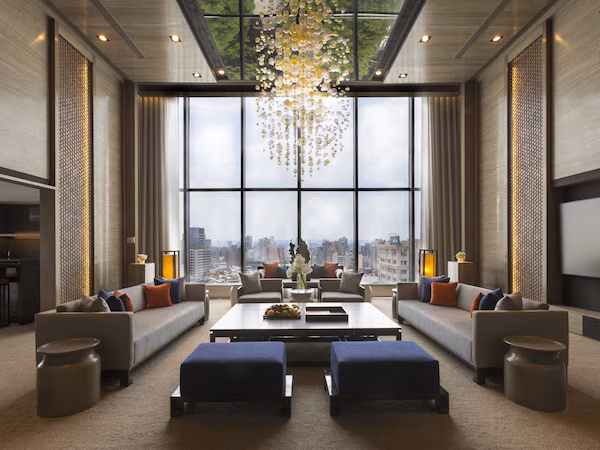 A spacious lounge area with a large window offering a city view, a modern chandelier, and a seating arrangement with sofas and ottomans.