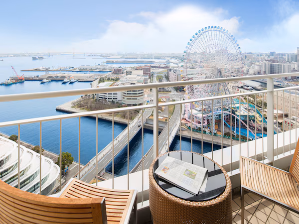 A balcony with wicker chairs, a small table, and a view of a waterfront with a Ferris wheel and amusement park.