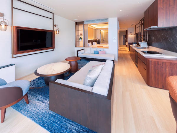 A modern hotel room with a sofa, a TV, a dining area, and a kitchen.