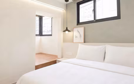 A bedroom with a bed, a bedside table, a nightstand, a wall-mounted light, a window, and a framed picture.