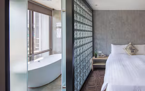 A modern hotel room with a large bathtub, a bed with white linens, and a decorative wall panel.