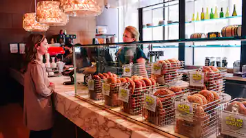 A bakery counter with various bagels and pastries displayed in wire baskets, a coffee machine, and a cash register.