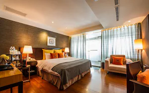A hotel room with a large bed, bedside tables, a desk, a chair, and a view of the outdoors through curtains.