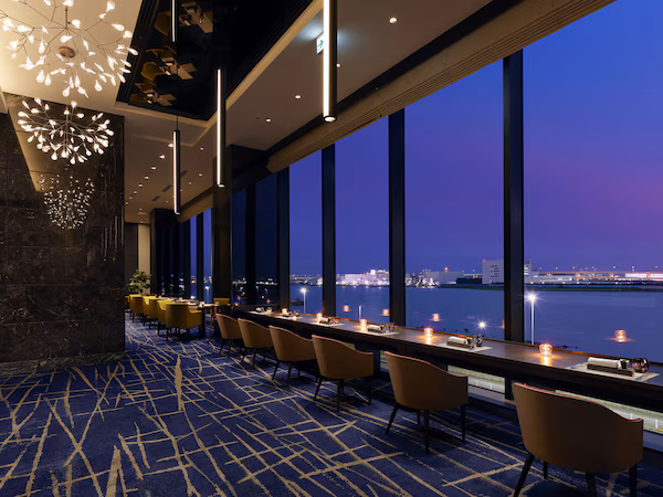 A modern hotel lobby with a long table, chairs, and a view of the water at dusk.