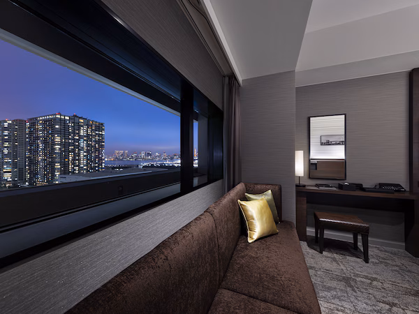 A modern hotel room with a large window offering a city view, a comfortable sofa, a desk with a chair, and a flat-screen TV.