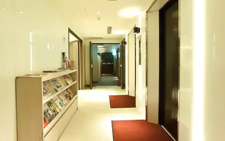 A hotel hallway with a reception desk, a bookshelf, and a door at the end.