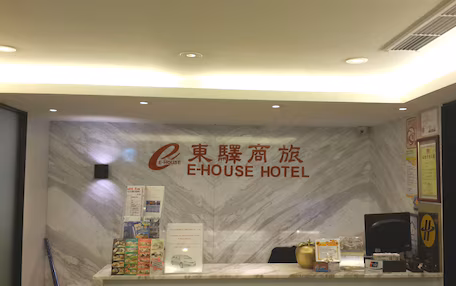 A reception desk with a marble wall, a sign reading "E-HOUSE HOTEL", and various informational brochures and a small display of items.