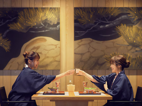 Two people seated at a table with a bottle and glasses, in a room with wooden paneling and a mural of pine trees.