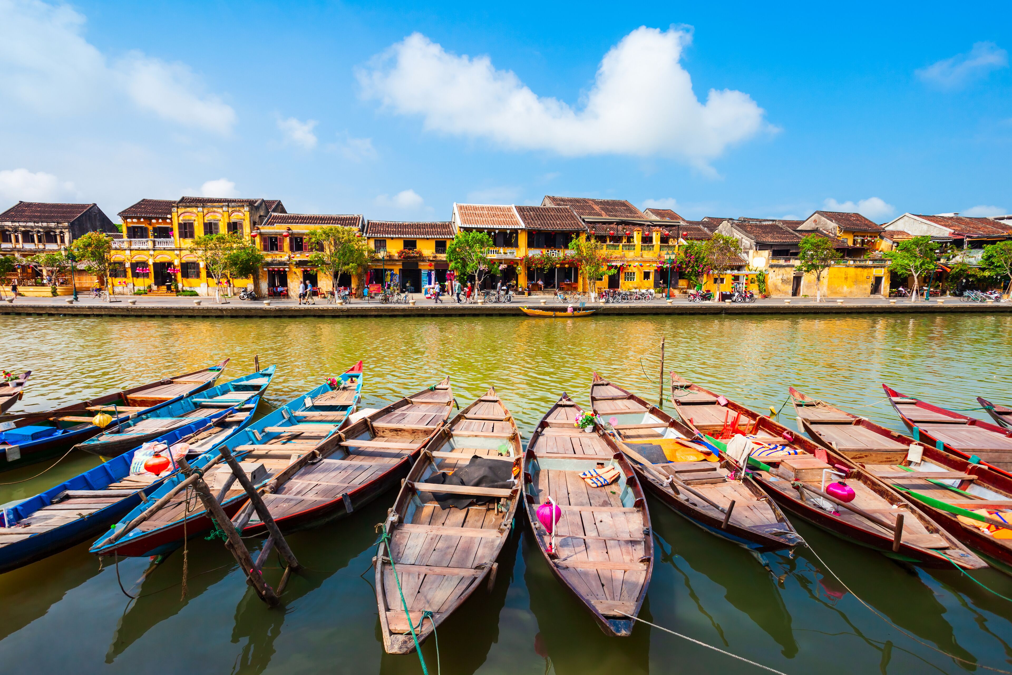 A row of colorful wooden boats docked along a riverbank with traditional yellow buildings in the background.