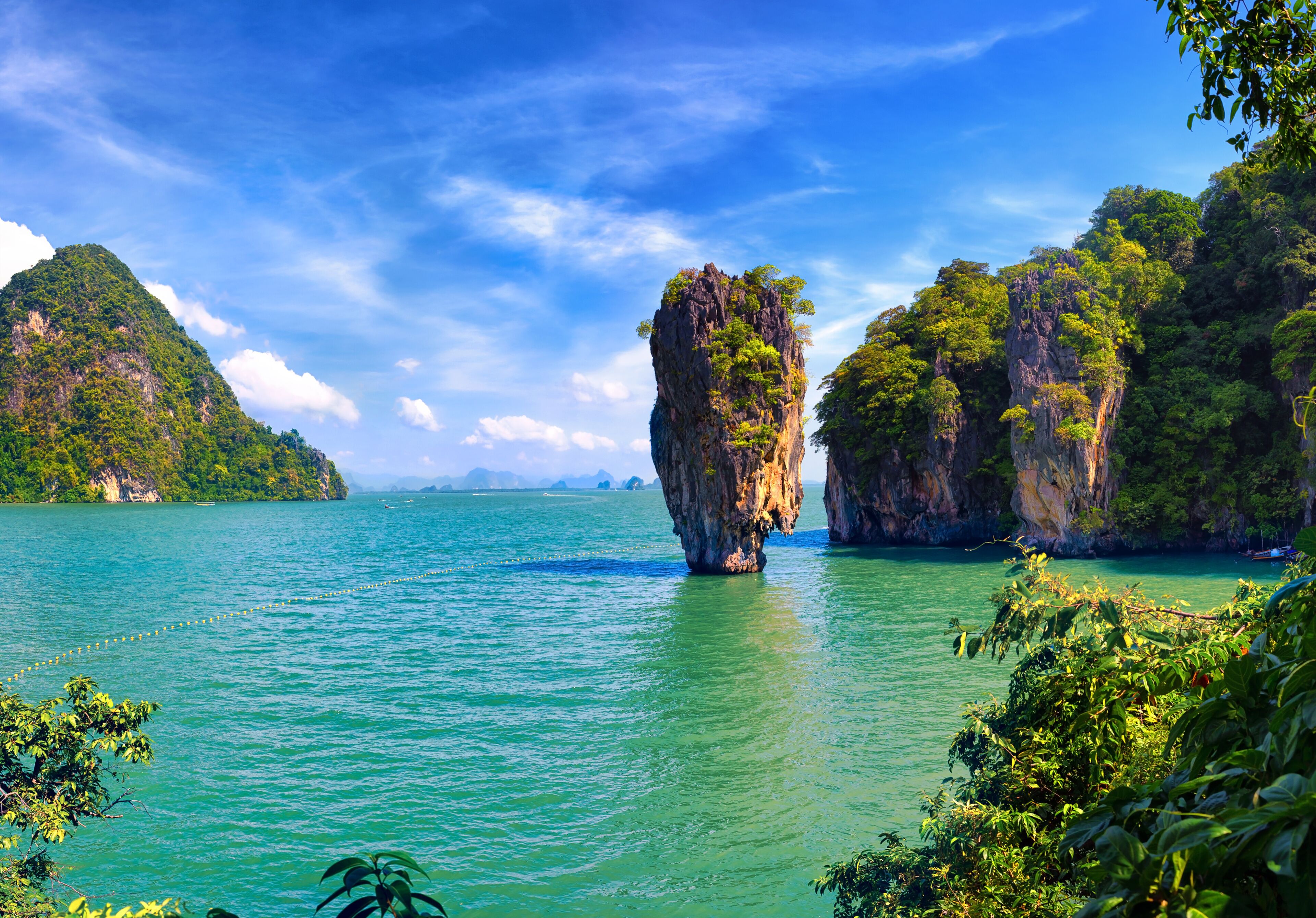 A clear turquoise sea with a prominent rock formation in the center, surrounded by lush green hills and a partly cloudy sky.